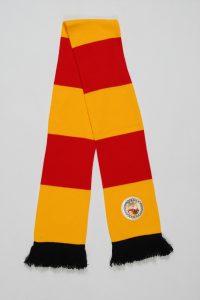 Ein Blockstreifenschal in Rot und Gelb mit schwarzen Fransen und einem Wappen.