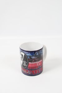 Kaffeebecher mit dem Logo des SC Freiburg und einem Stadionmotiv in rot und blau.