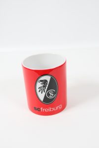 Roter Kaffeebecher mit dem Logo des SC Freiburg auf der Vorderseite.