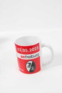 Kaffeebecher mit dem Logo von SC Freiburg und dem Datum 02.05.2023 auf rotem Hintergrund.
