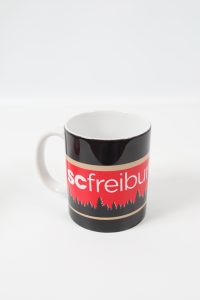 Schwarzer Kaffeebecher mit rotem SC Freiburg Logo und Baumgrafik.