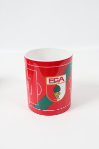 Roter Kaffeebecher mit dem Logo des FC Augsburg und einem Fußballfeld-Design.