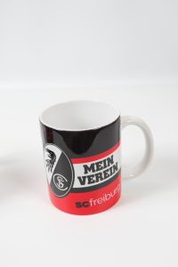 Schwarz-rot-weißer Kaffeebecher mit dem Logo von SC Freiburg und dem Text 'Mein Verein'.