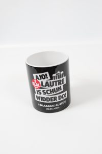 Schwarzer Kaffeebecher mit verschiedenen Texten in Weiß und Rot, darunter 'AJO!' und 'IS SCHUN WIDDER DO!'.