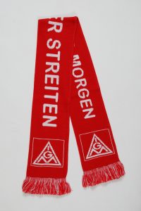 Roter Strickschal mit weißen Schriftzügen und Fransen, zeigt ein geometrisches Logo.