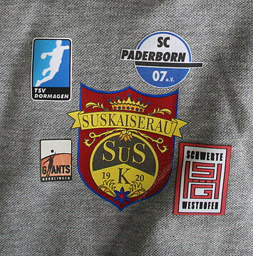 Wappen von Sus Kaiserau mit Logos von SC Paderborn und TSV Dormagen auf einem grauen Hintergrund.