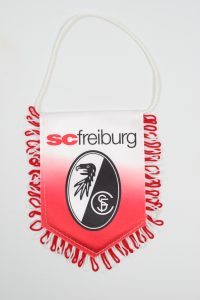 Wimpel des SC Freiburg mit dem Vereinslogo und roten Fransen, auf weißem Hintergrund.