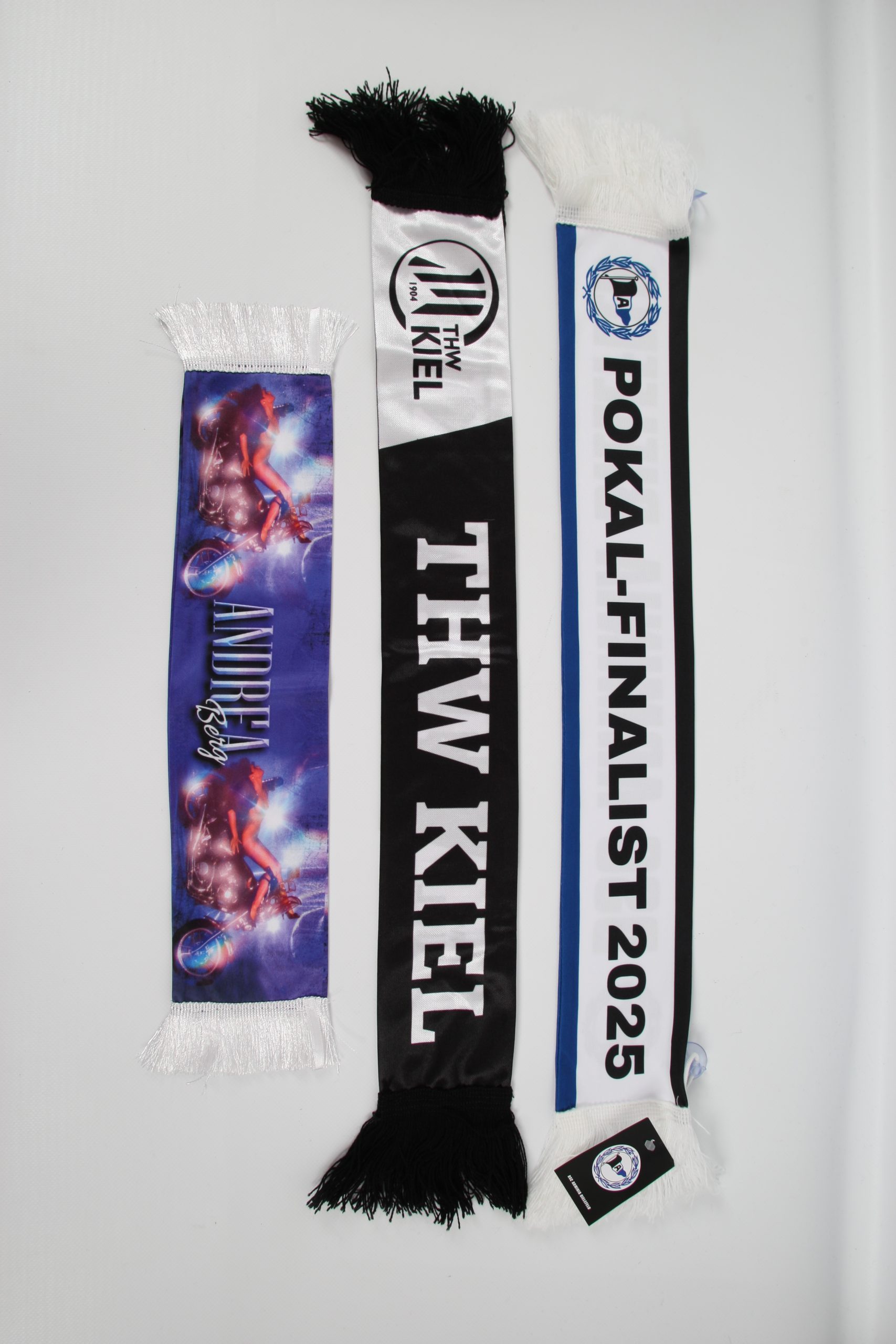 Drei Schals, darunter ein THW Kiel Schal und ein Schal für Pokal-Finalist 2025, mit Fransen an den Enden.