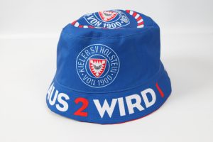 Blauer Fischerhut mit dem Logo des Kieler Sportvereins von 1900 und dem Schriftzug 'AUS 2 WIRD 1'.