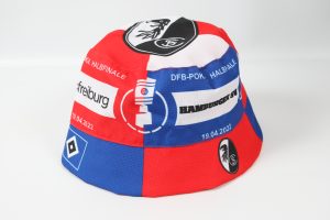 Bunter Fischerhut mit verschiedenen Vereinslogos in Rot, Blau und Weiß, einschließlich SC Freiburg und Hamburger SV.