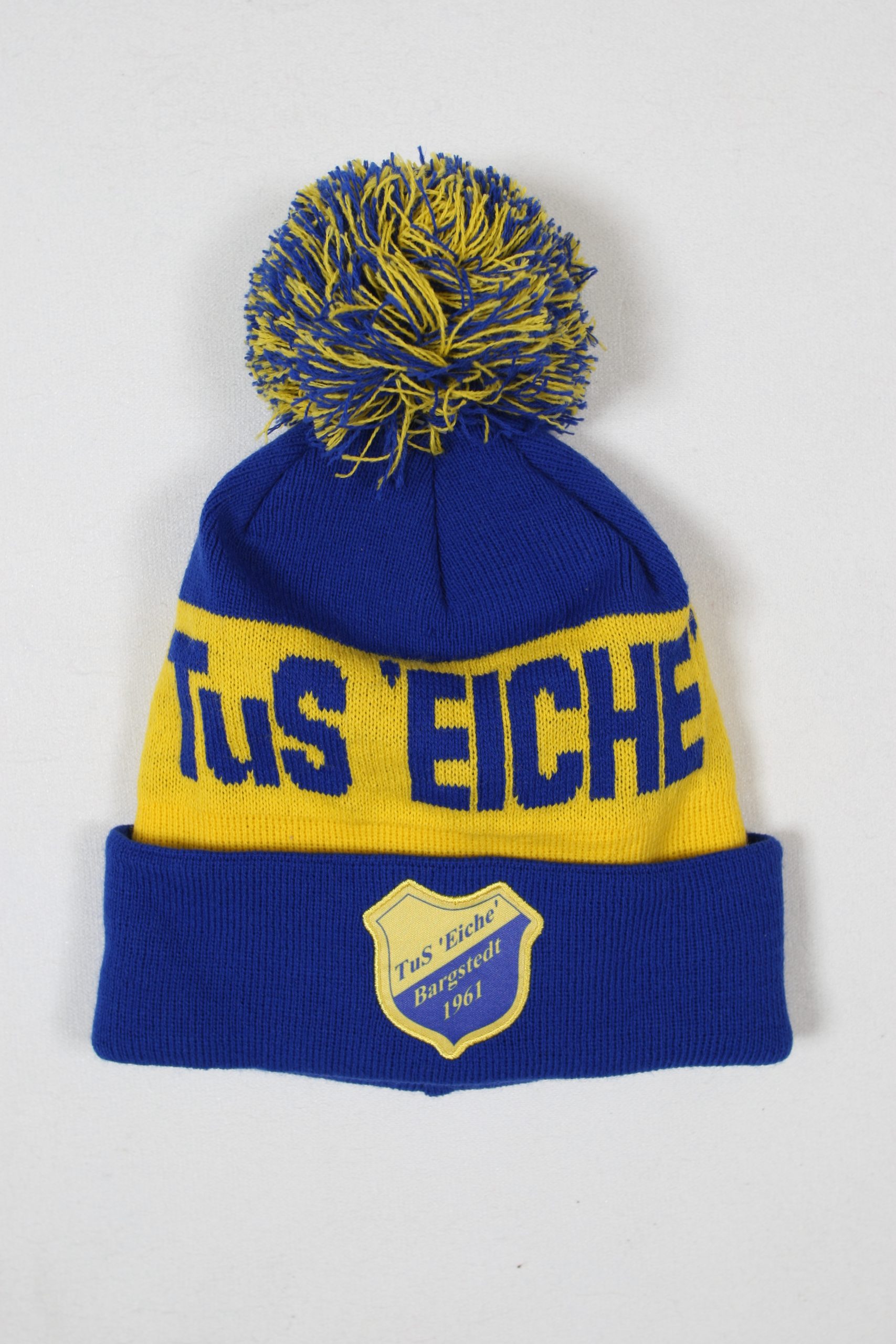 Blaue und gelbe Mütze mit einem Bommel und dem Vereinslogo von TuS Eiche Harsfeld 1961.