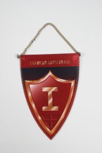 Roter Wimpel mit goldener Schrift und einem Emblem in der Mitte, an einer Kette aufgehängt.