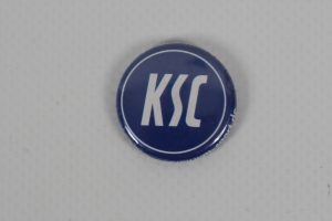 Blauer Button mit dem KSC-Logo in weißer Schrift auf einem neutralen Hintergrund.