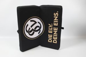 Schwarzes Klappkissen mit einem Logo und dem Schriftzug 'DIE ELY! DEINE EINS.' auf der Vorderseite.