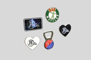 Eine Sammlung von Magneten mit den Logos von THW Kiel und HSG Wetzlar, darunter ein Herzmagnet und ein Flaschenöffner.