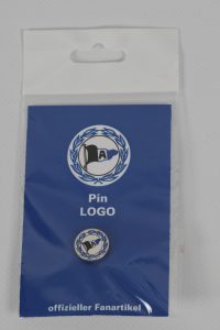 Pin mit Logo in einer transparenten Verpackung auf blauem Hintergrund.