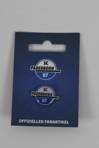 Zwei Anstecknadeln mit dem Logo des SC Paderborn 07 auf einer blauen Karte.