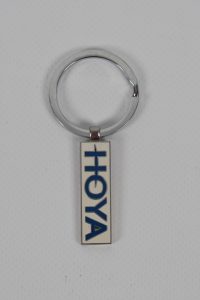 Metallischer Schlüsselanhänger mit dem Hoya Logo in blauer Schrift auf weißem Hintergrund.