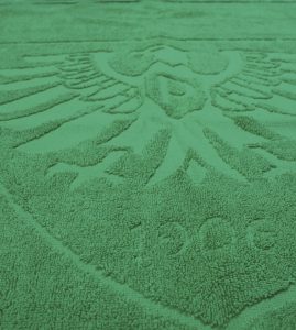 Ein grün gefärbtes Frottierhandtuch zeigt eine geprägte Bordüre mit feinem Strukturmuster und verweist auf die bedruckte Ausführung aus 100 % Baumwolle.