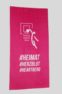 Ein pinkes Frottierhandtuch von Telekom Baskets Bonn zeigt das Logo und die Hashtags #HEIMAT, #HERZBLUT und #HEARTBERG im weißen Druck.