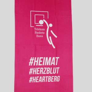 Ein pinkes Frottierhandtuch von Telekom Baskets Bonn zeigt das Logo und die Hashtags #HEIMAT, #HERZBLUT und #HEARTBERG im weißen Druck.