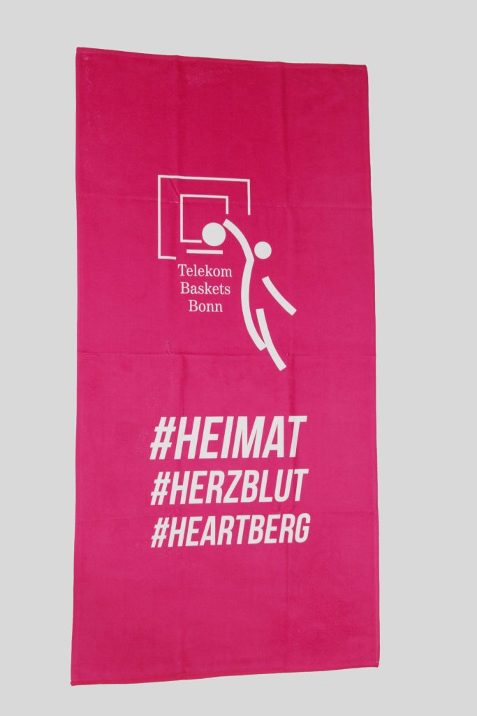 Ein pinkes Frottierhandtuch von Telekom Baskets Bonn zeigt das Logo und die Hashtags #HEIMAT, #HERZBLUT und #HEARTBERG im weißen Druck.
